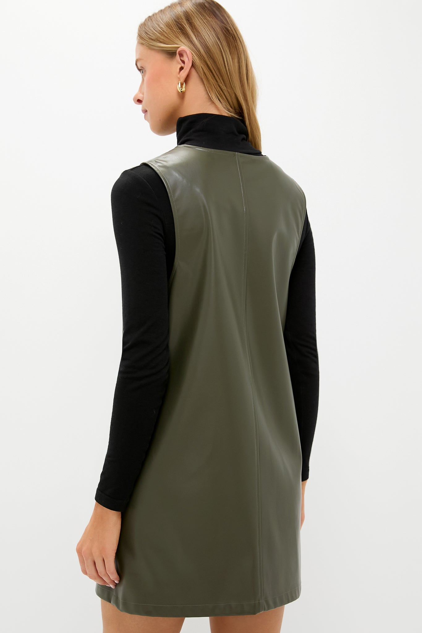 Woman in an olive faux leather pinafore dress over a black turtleneck, embodying a classic, effortless style with a single gold hoop earring.