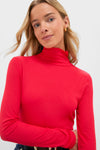 A person with long blonde hair wears a bright red Tuckernuck Lightweight Ribbed Turtleneck in a slim fit and gold hoop earrings, standing against a plain light background.