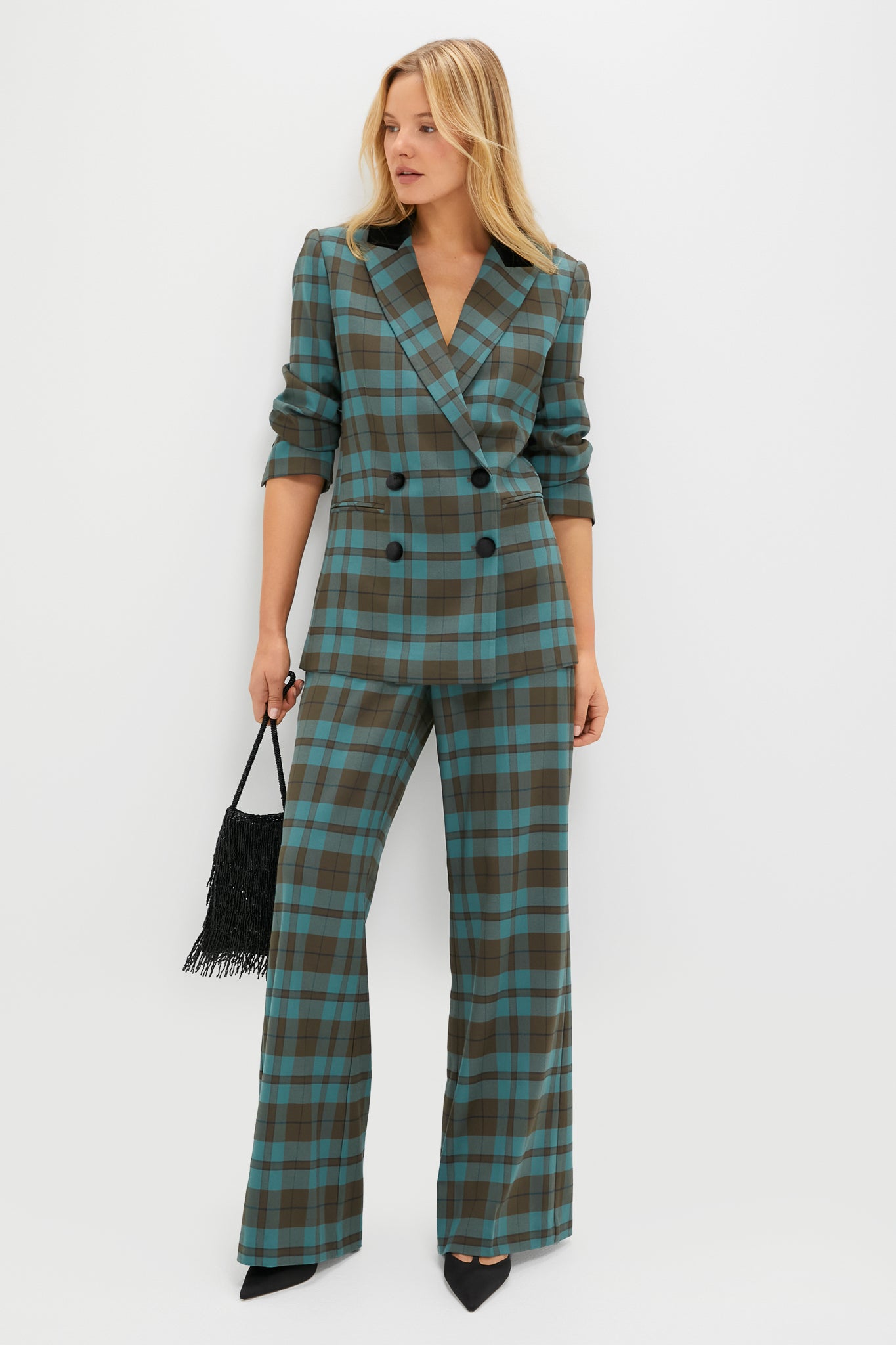 A woman stands confidently against a white background wearing a Tuckernuck teal and brown plaid blazer with wide-leg pants, paired with black pointed-toe shoes and a black fringe handbag.