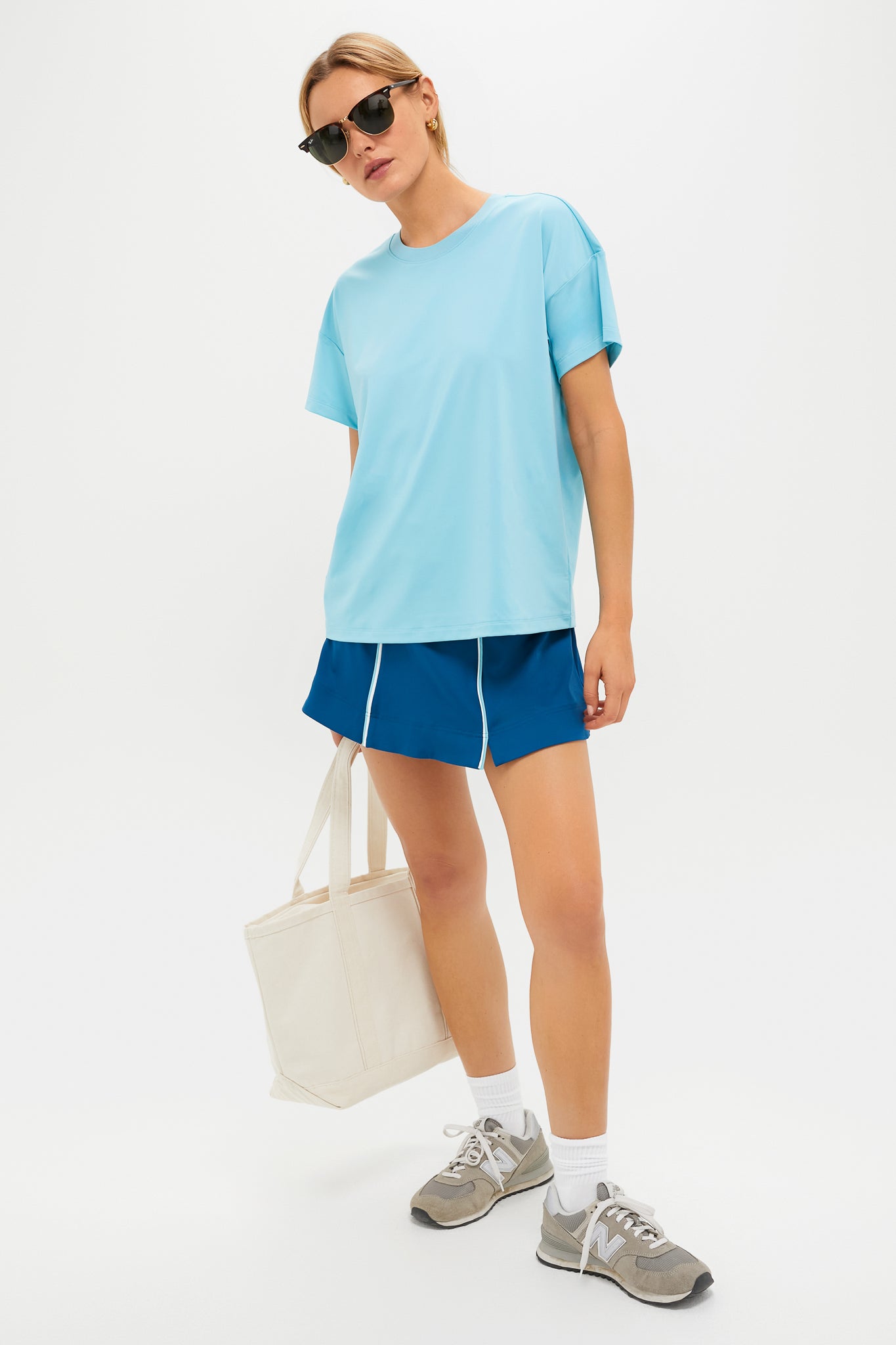 Wearing a Tnuck Sport light blue crewneck t-shirt, blue shorts, gray sneakers, and sunglasses, this person holds a beige tote bag while standing against a plain white background.