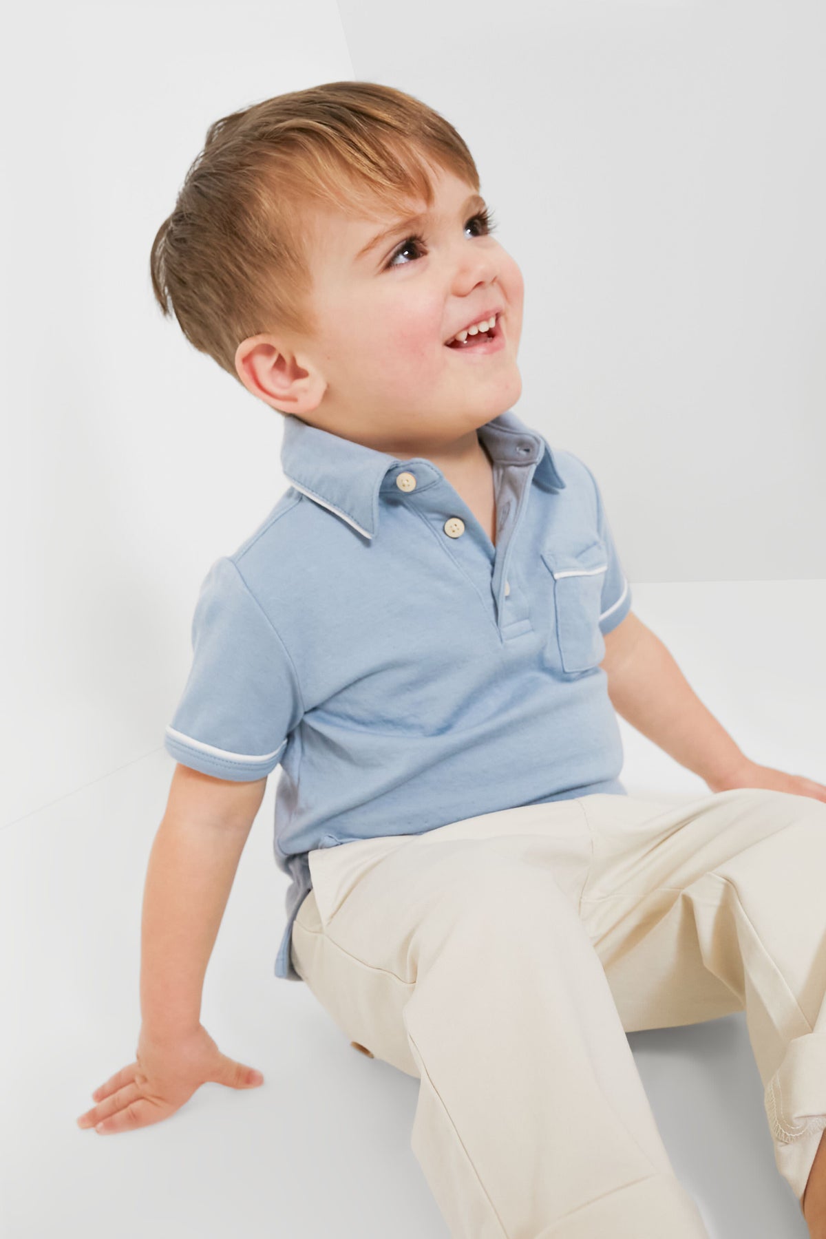 A young boy wearing an Oso & Me light blue unisex pima cotton polo and beige pants sits on the floor, looking up with a smile.