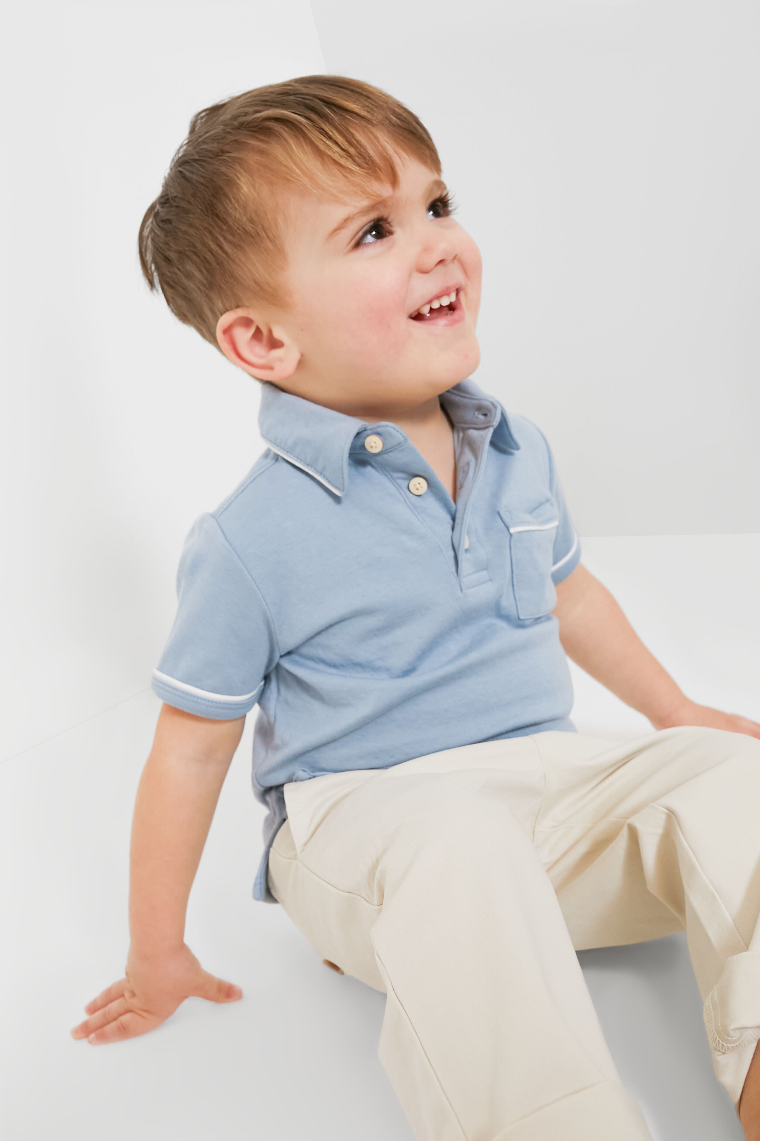 A young boy wearing an Oso & Me light blue unisex pima cotton polo and beige pants sits on the floor, looking up with a smile.