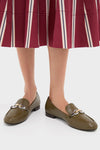 A person wears Tory Burch moss-green loafers with metal chain details, paired with a pleated maroon skirt featuring vertical stripes.