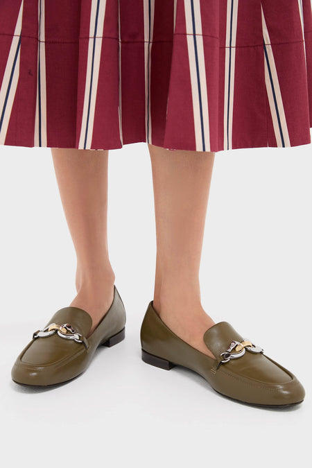 A person wears Tory Burch moss-green loafers with metal chain details, paired with a pleated maroon skirt featuring vertical stripes.