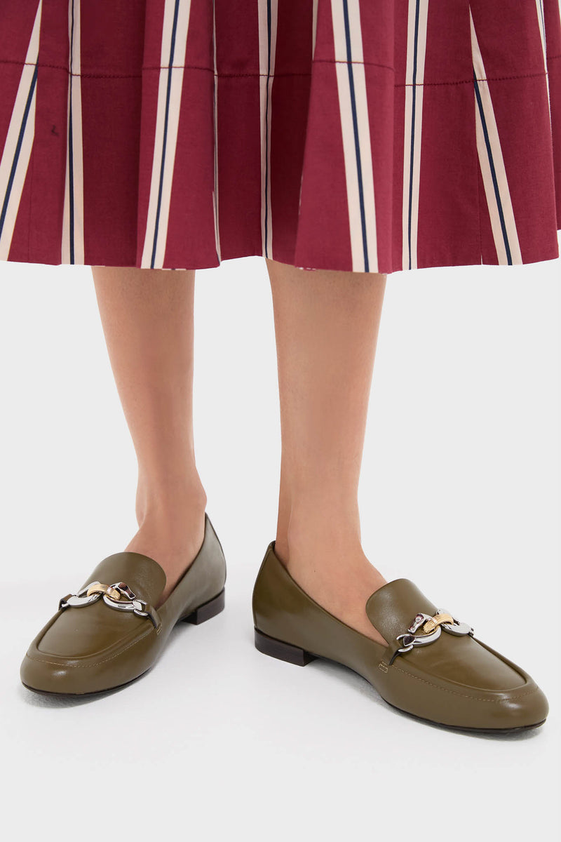 A person wears Tory Burch moss-green loafers with metal chain details, paired with a pleated maroon skirt featuring vertical stripes.