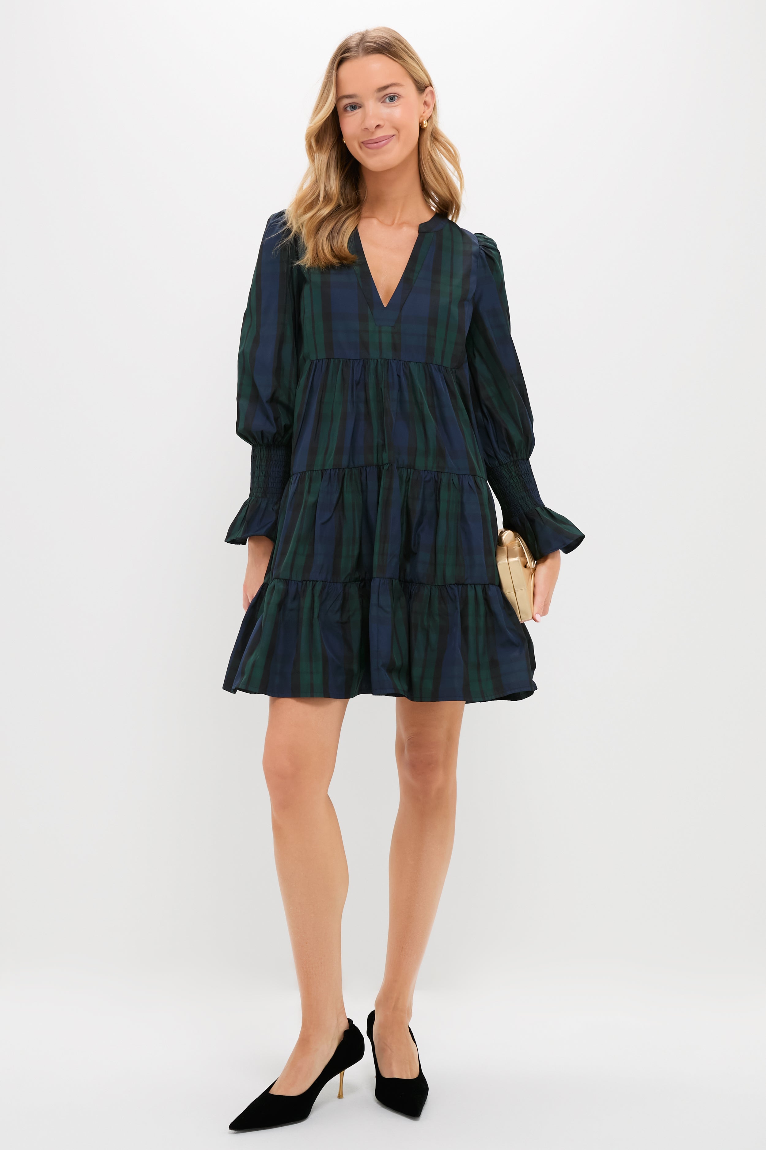 Woman against a plain background wears a tiered, long-sleeve Pomander Place Blackwatch dress with V-neckline, black heels, and a beige clutch—an effortlessly chic holiday outfit.