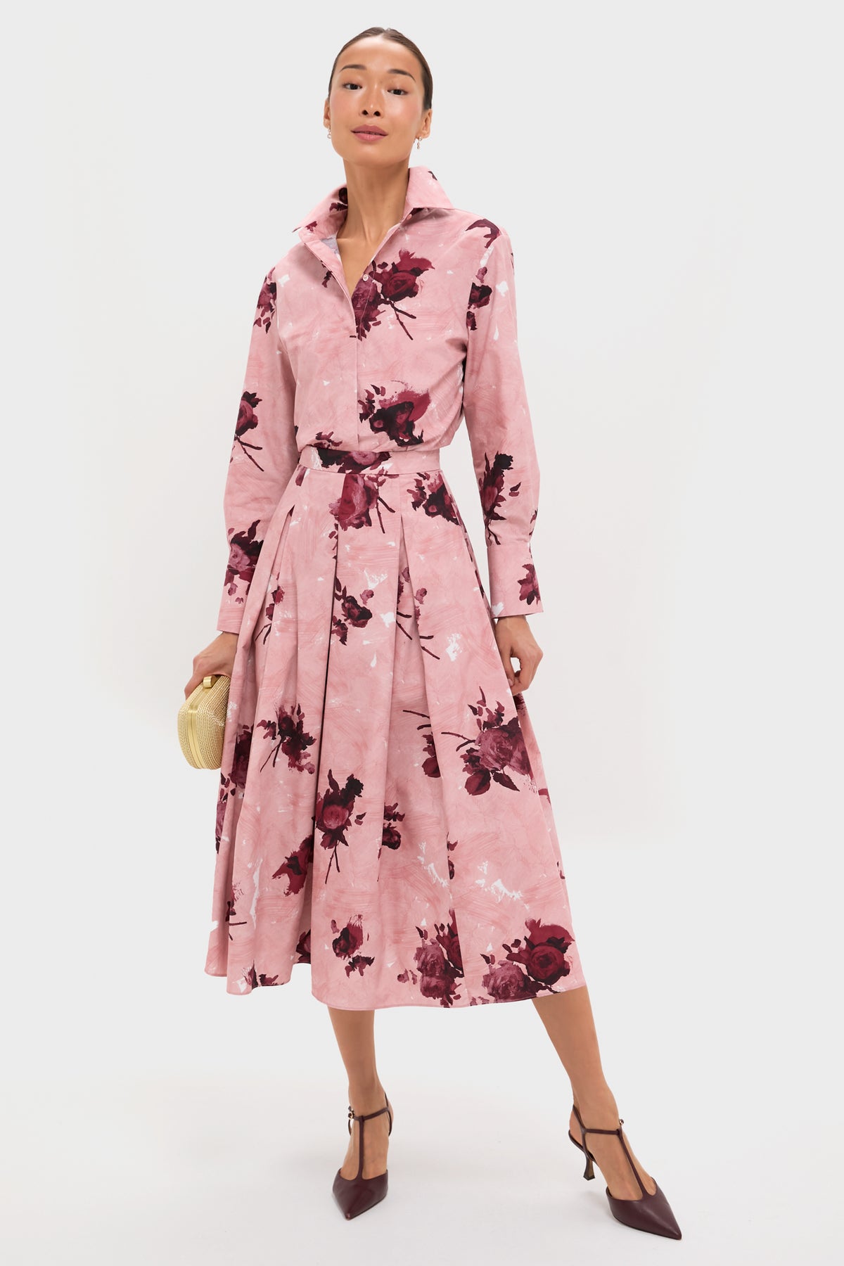 A woman wears an ERDEM pink, long-sleeve collared midi dress with dark floral motifs, holding a beige clutch and pairing it with pointed burgundy heels.