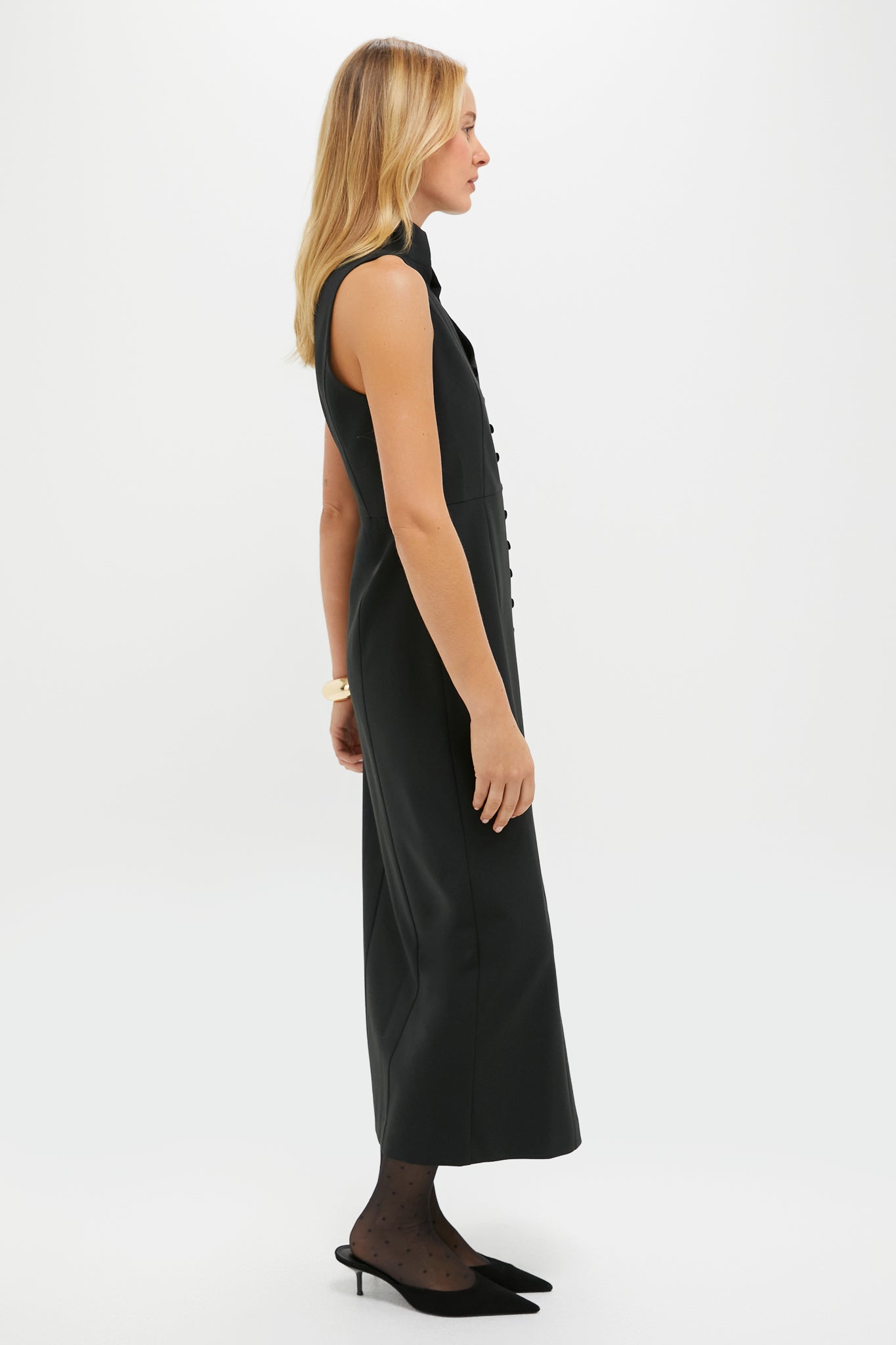 A woman stands in profile against a plain white background, wearing a Tuckernuck sleeveless velvet dress, black sheer polka-dot tights, and black pointed heels.