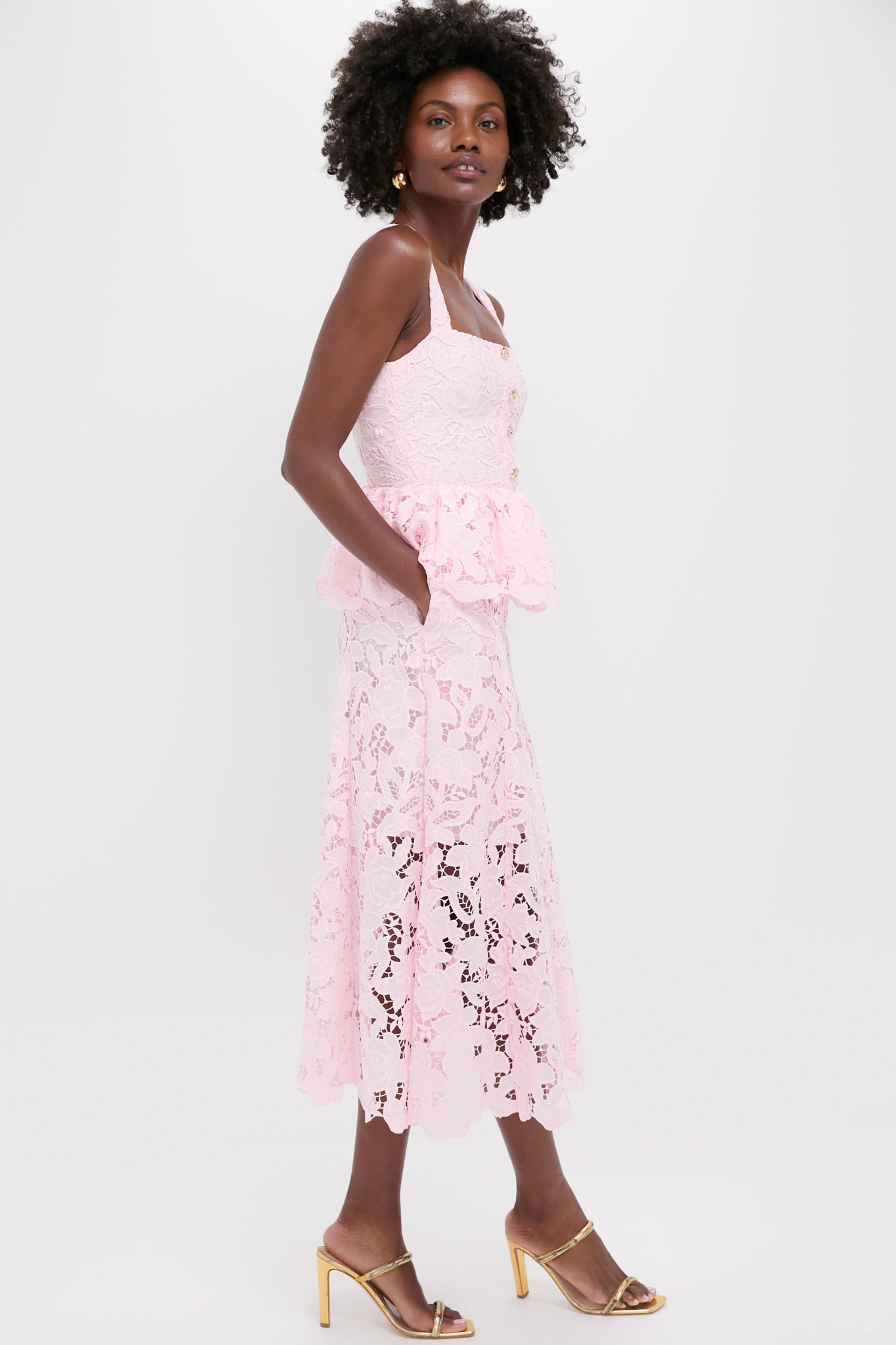 A woman in profile wears a Self-Portrait romantic lace peplum dress with spaghetti straps and a sweetheart neckline, styled with gold heeled sandals, set against a plain white background.