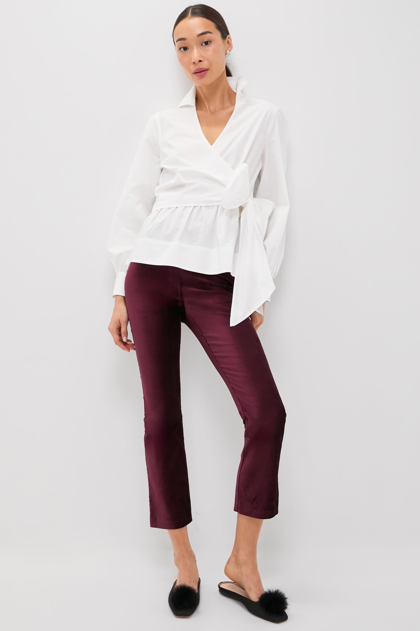 A woman stands against a white background wearing a Tuckernuck white wrap blouse, high rise burgundy velvet kick flare pants, and black fur mule slippers.