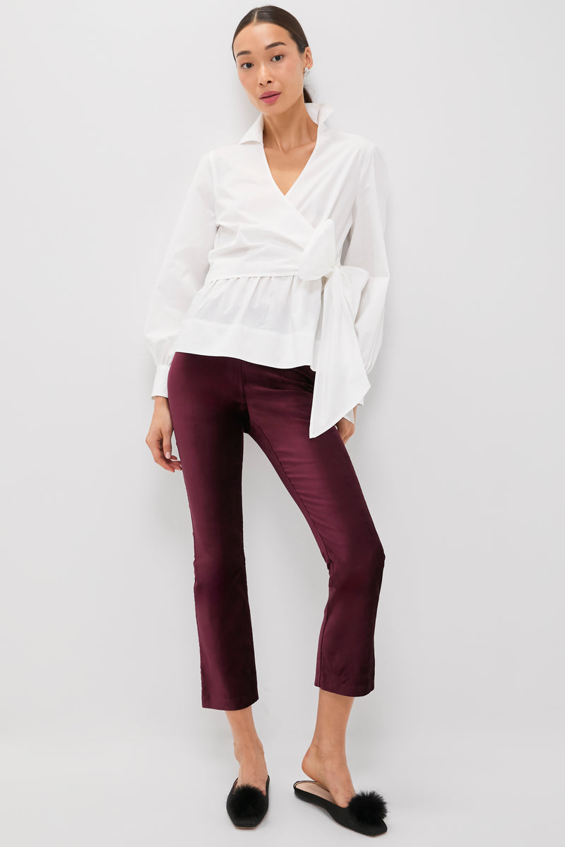 A woman stands against a white background wearing a Tuckernuck white wrap blouse, high rise burgundy velvet kick flare pants, and black fur mule slippers.