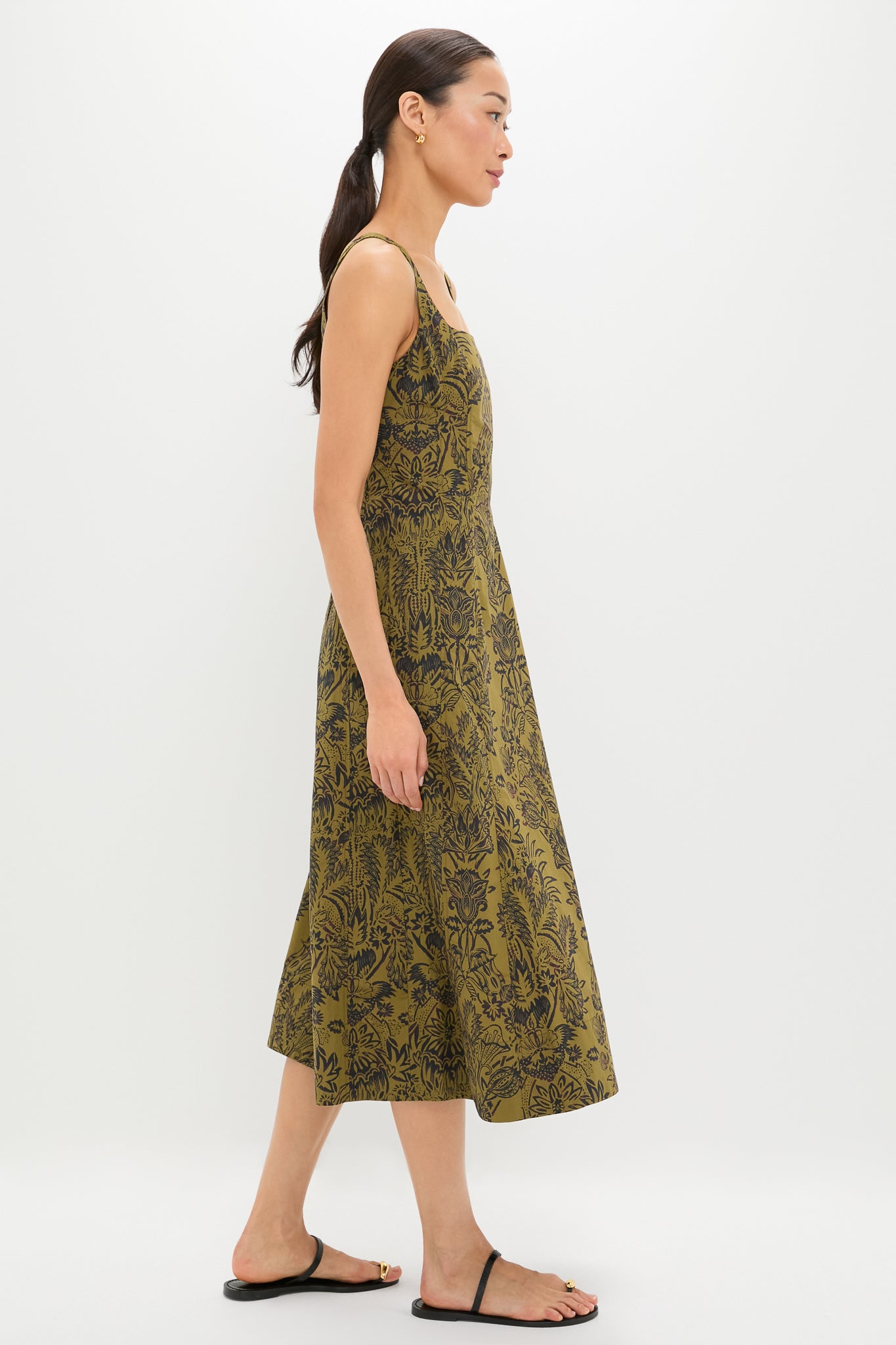 Woman wearing the Olive Flora Natali Dress, a sleeveless midi with an all-over black botanical print, embodies classic, effortless style. Paired with black sandals and gold hoops for a high-end look.