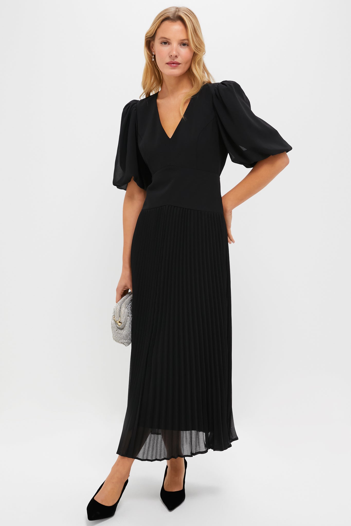 A woman wears a pleated midi dress with puff sleeves and a V-neck, styled with a silver clutch and black heels.