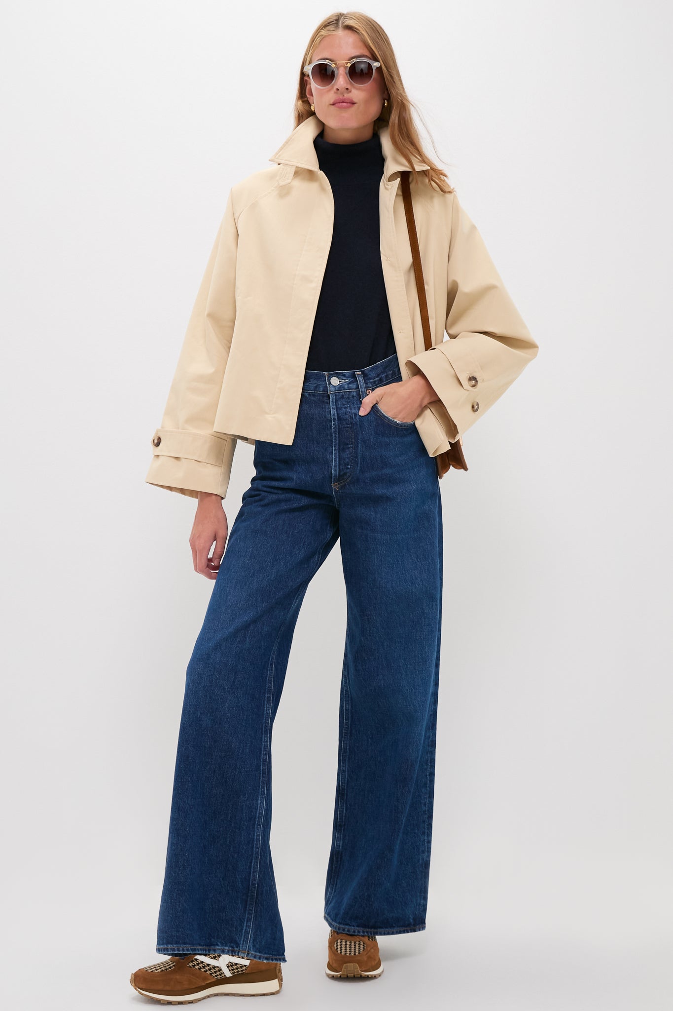 A woman wears a beige jacket, black turtleneck, and wide-leg dark blue jeans, styled with brown shoes and sunglasses.