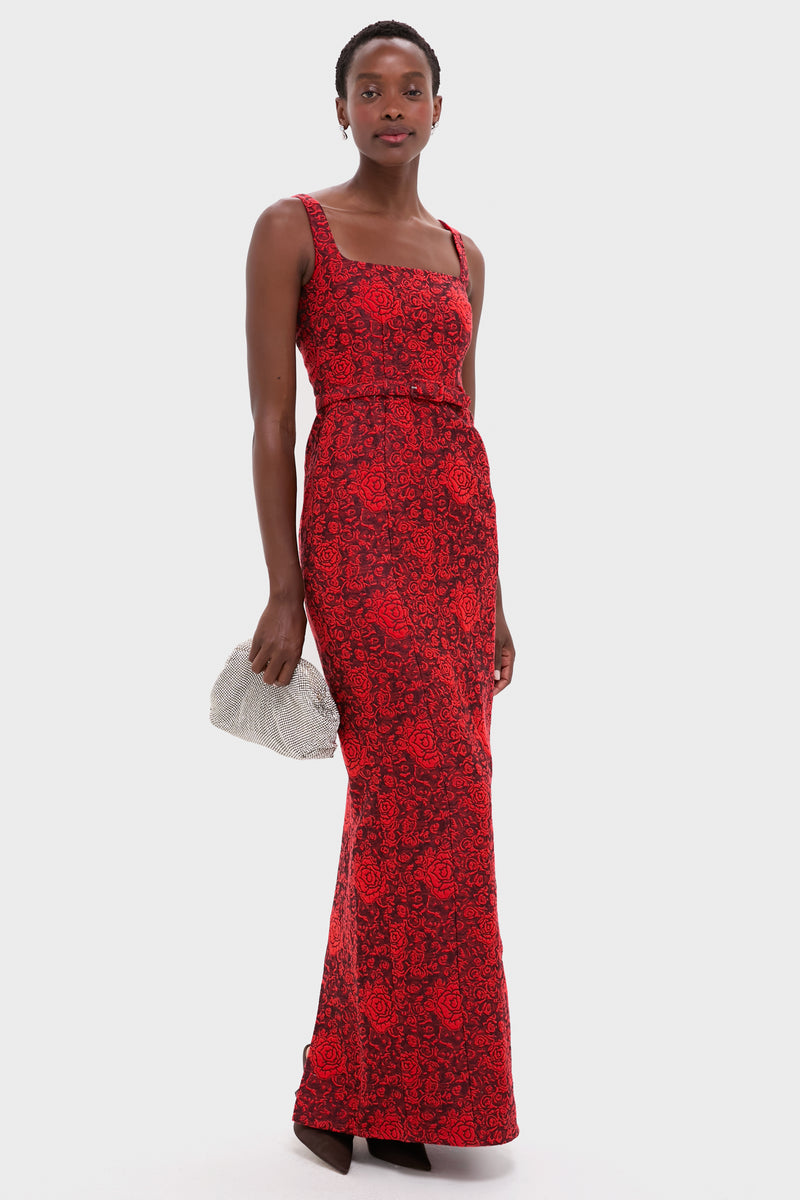 A woman wears a sleeveless, floor-length Saloni Bora Long Dress in red Rose Brocade featuring a black floral pattern, holds a silver clutch, and stands against a plain white background.