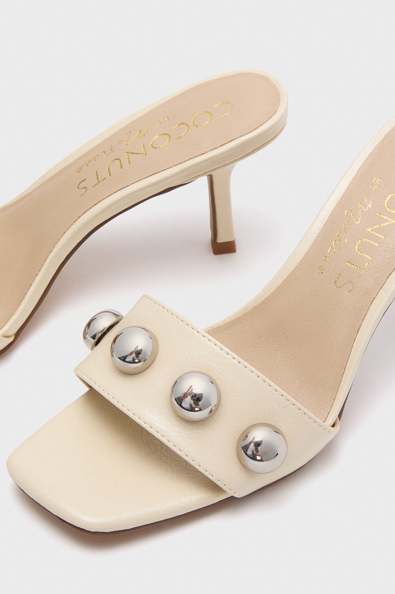 Matisse cream heeled mules feature open toes, square fronts, and wide front straps adorned with metallic round studs.