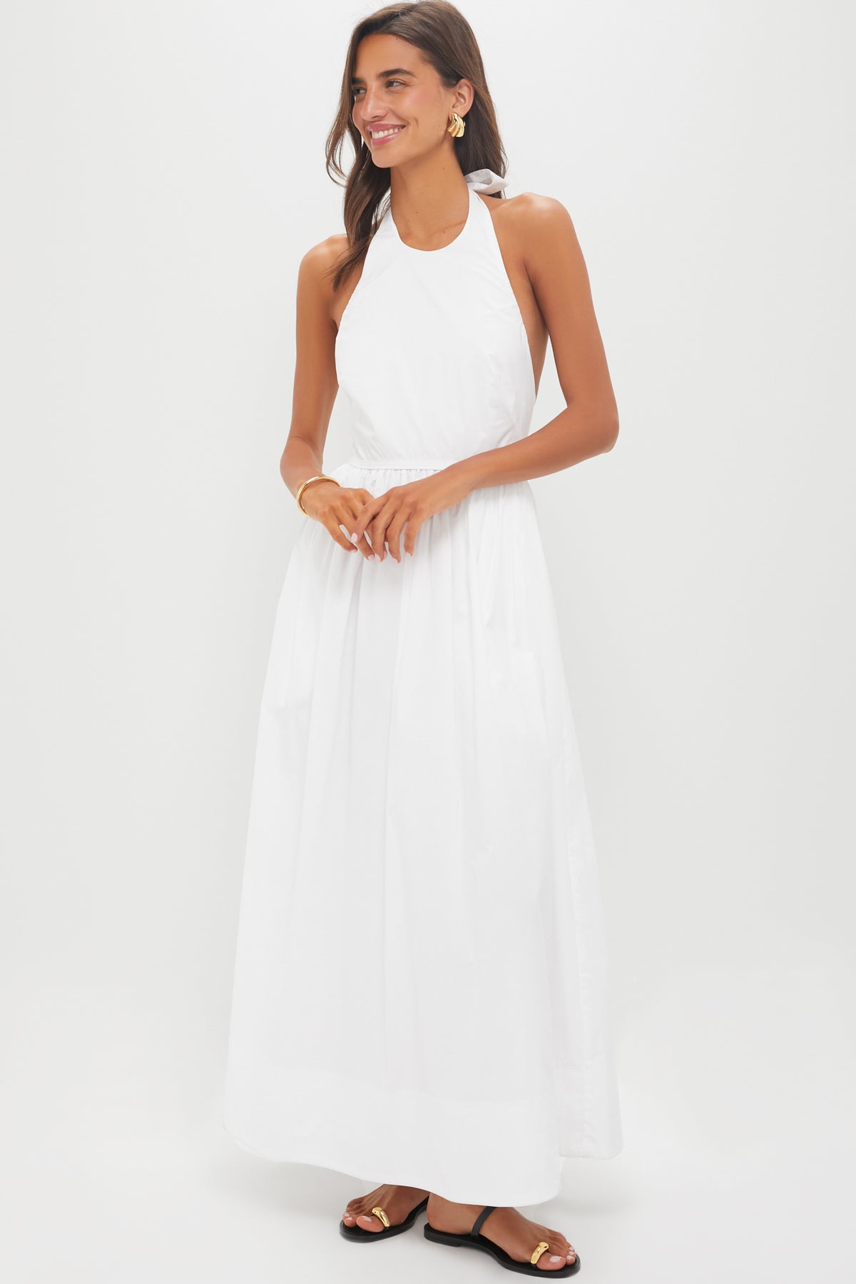 A woman wears a sleeveless, halter neck white Tuckernuck summer dress (100% cotton, cinched waist), gold hoop earrings, a gold bracelet, and black flat sandals—perfect for warm weather and effortless casual elegance.