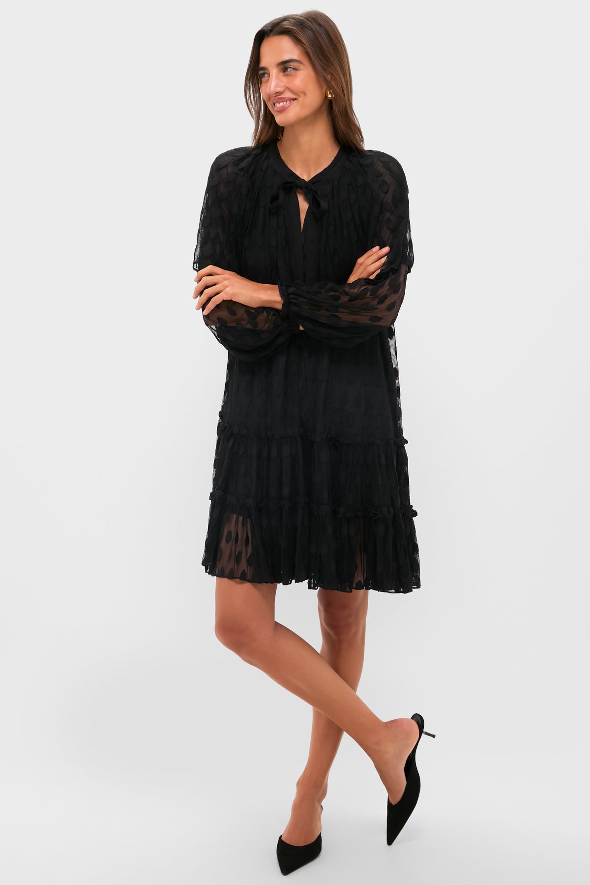 A woman stands with arms crossed in a black Ulla Johnson tiered dress with a tie neckline and black pointed-toe heels against a plain light background.