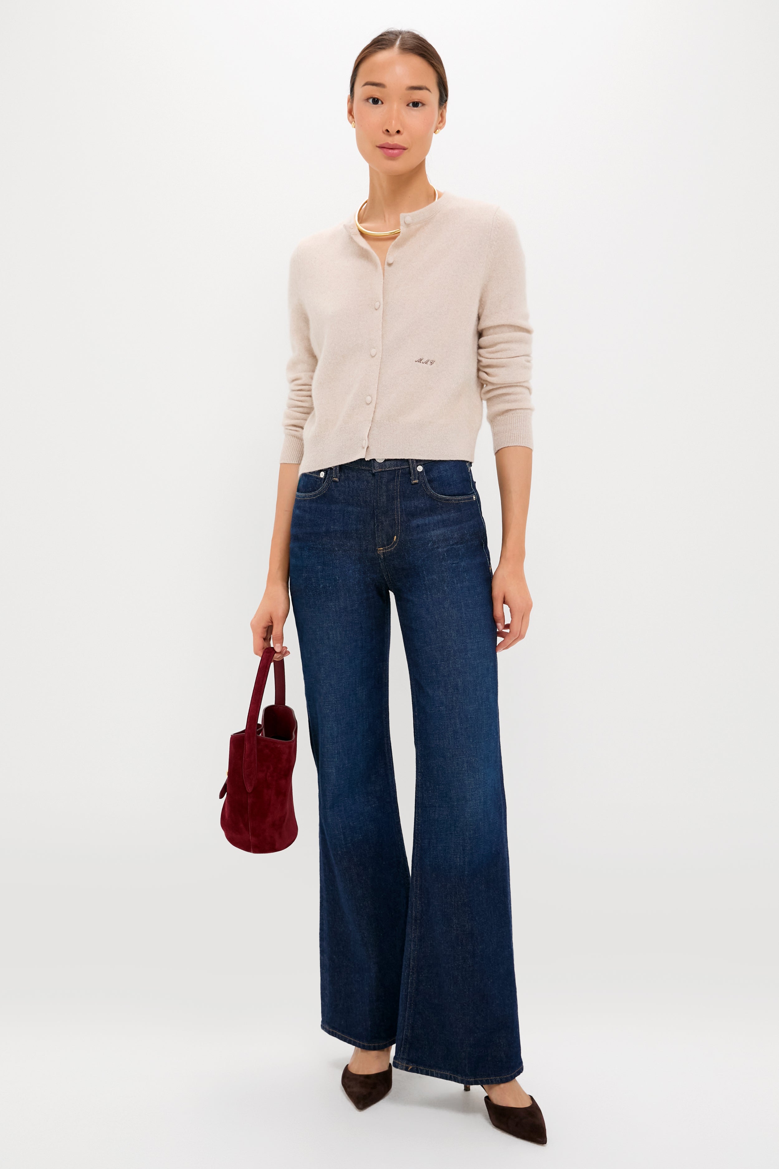 A woman wears a Tuckernuck beige cashmere cardigan, dark blue wide-leg jeans, brown pointed shoes, and holds a small red bag against a plain white background.