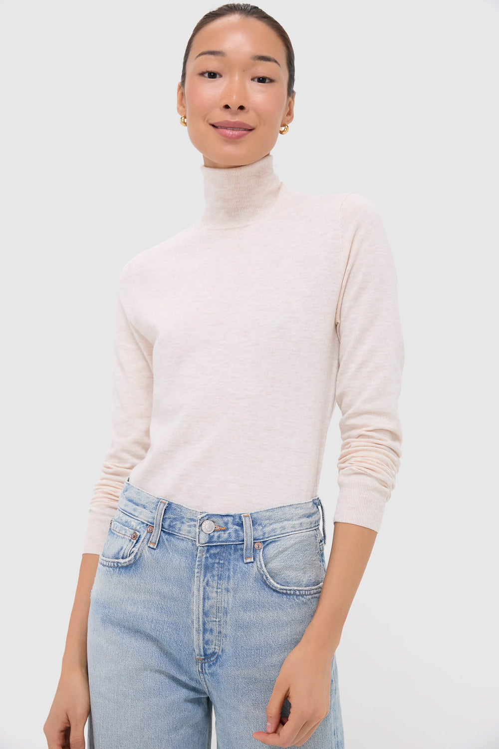 A person wears a Nude Lucy cream turtleneck and light blue high-waisted jeans, effortlessly showcasing style in a Nude Lucy light beige merino wool sweater.