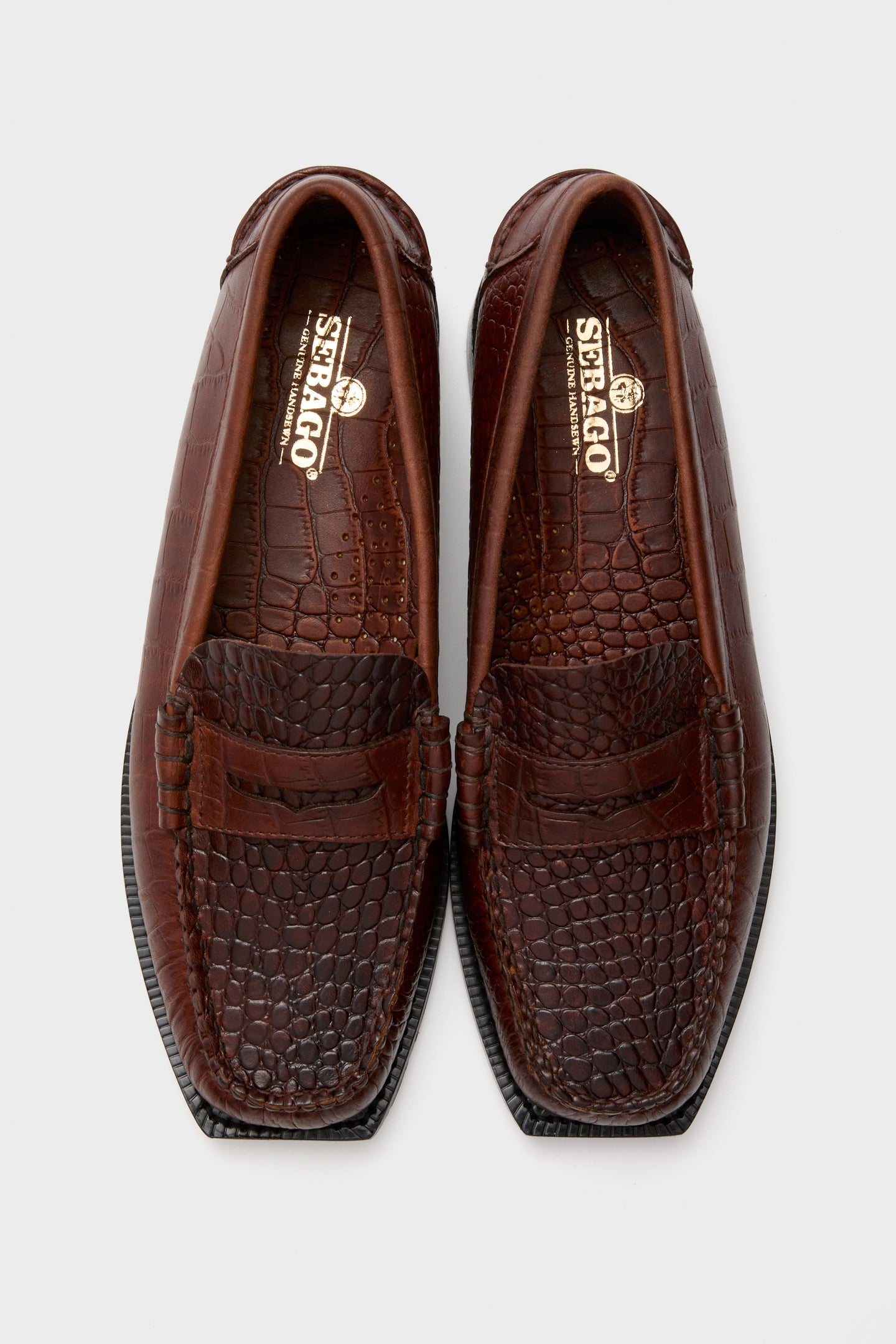 A pair of SEBAGO brown crocodile-patterned leather loafers, featuring the brand’s logo on the insole, shown from above on a white background.