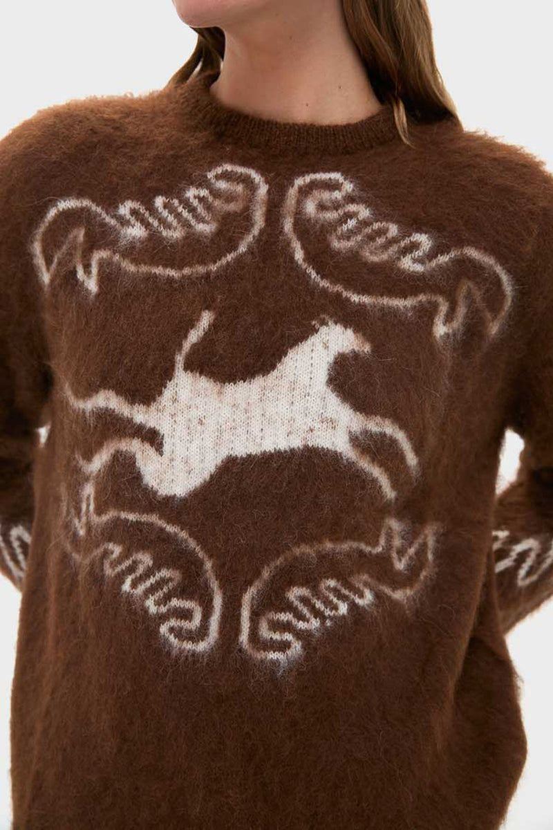 A person wears a fuzzy brown Carolina K alpaca knit sweater featuring a white abstract horse and swirl pattern on the front and sleeves, shown from the shoulders down.