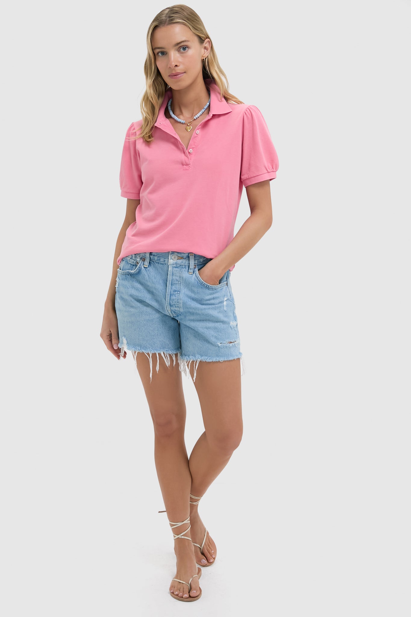 A woman wears a pink Tuckernuck Peruvian Cotton pique polo shirt, light blue frayed denim shorts, and strappy sandals.