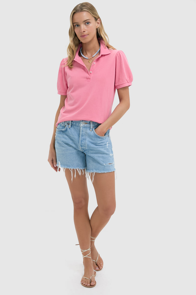 A woman wears a pink Tuckernuck Peruvian Cotton pique polo shirt, light blue frayed denim shorts, and strappy sandals.