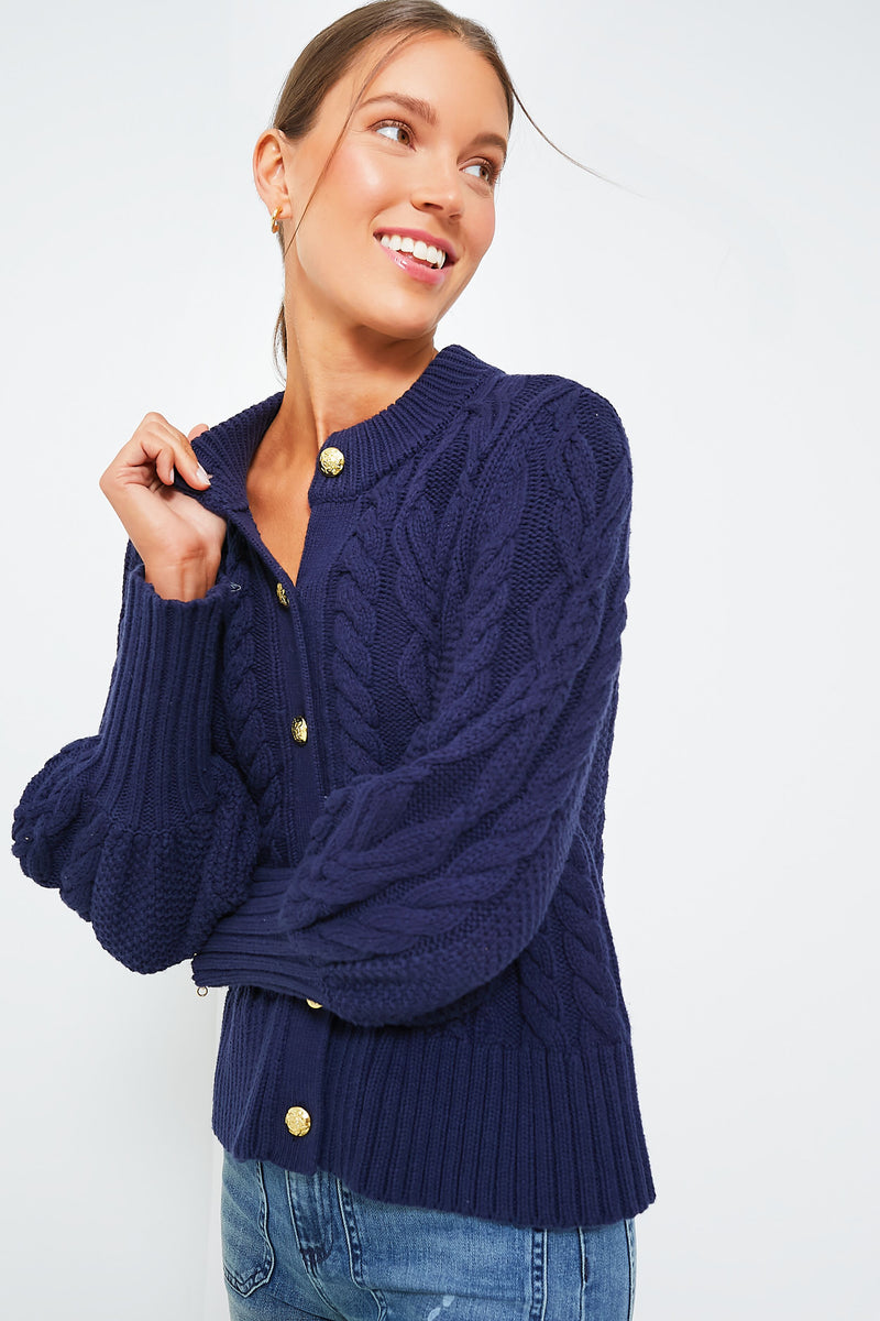 Woman wearing the Tuckernuck Navy Colette Cardigan, a chunky cable-knit style with gold buttons, paired with blue jeans and standing against a plain white background.