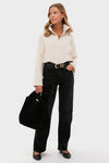 A woman stands against a plain background wearing a Pomander Place cream long sleeve polo with a relaxed fit, black jeans with a belt, black flats, and holding a black bag.