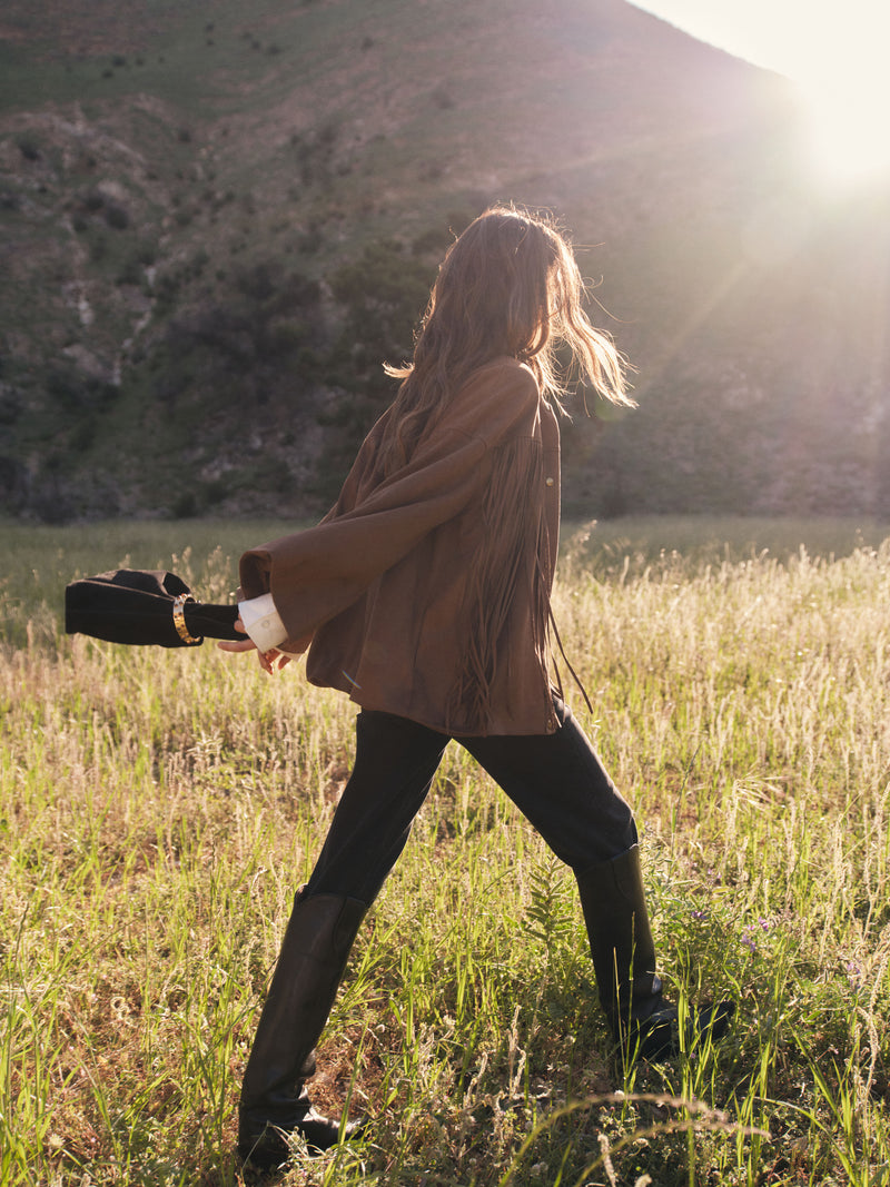 Woman walking away from the camera in a brown fringe jacket, pants, and black leather boots. She's swinging a brown bag with a gold detail behind her. 