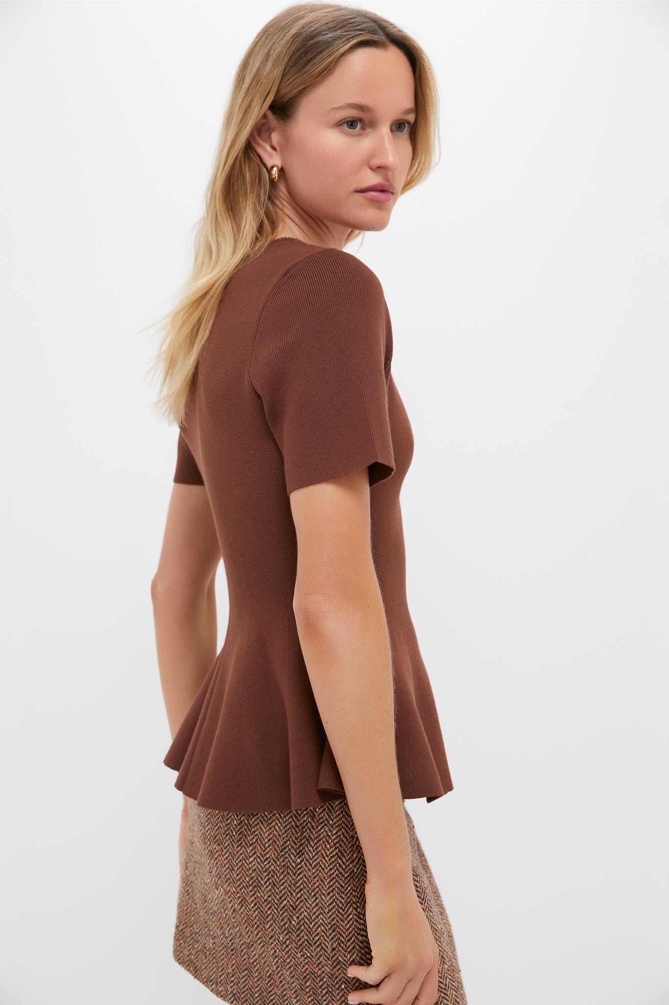 Woman in classic, effortless Chocolate Brown Knit Phoebe Peplum Top with playful peplum hem, embodying fun, high-end style, paired with tweed-patterned skirt, gold hoop earrings.