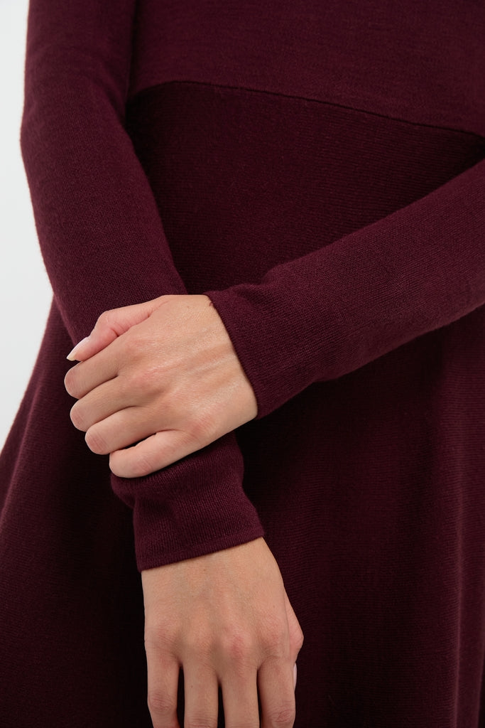 A person wears a Pomander Place long-sleeve burgundy sweater dress, standing with one hand holding the opposite wrist against a plain background.
