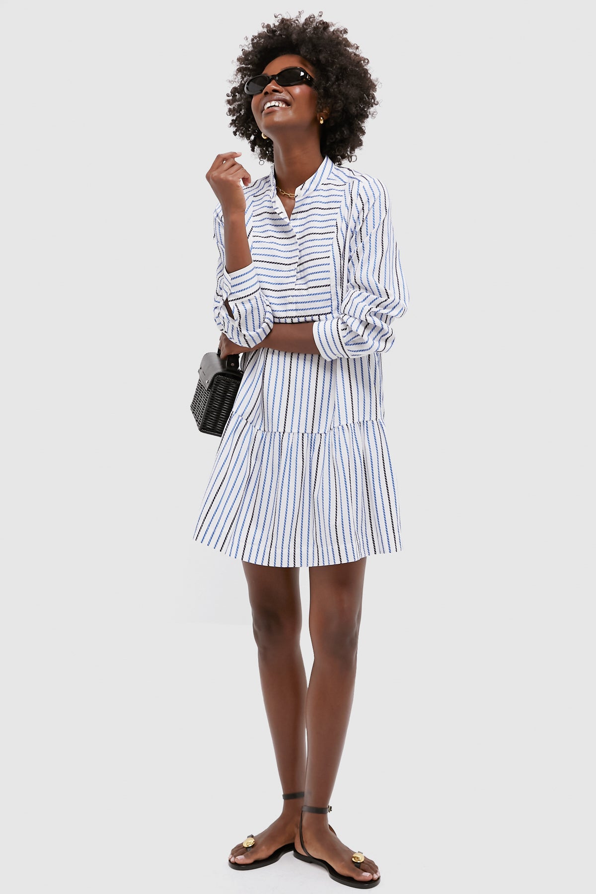 A woman in a classic Tuckernuck blue and white striped mini dress, black sandals, sunglasses, and a small black purse stands against a plain light background.