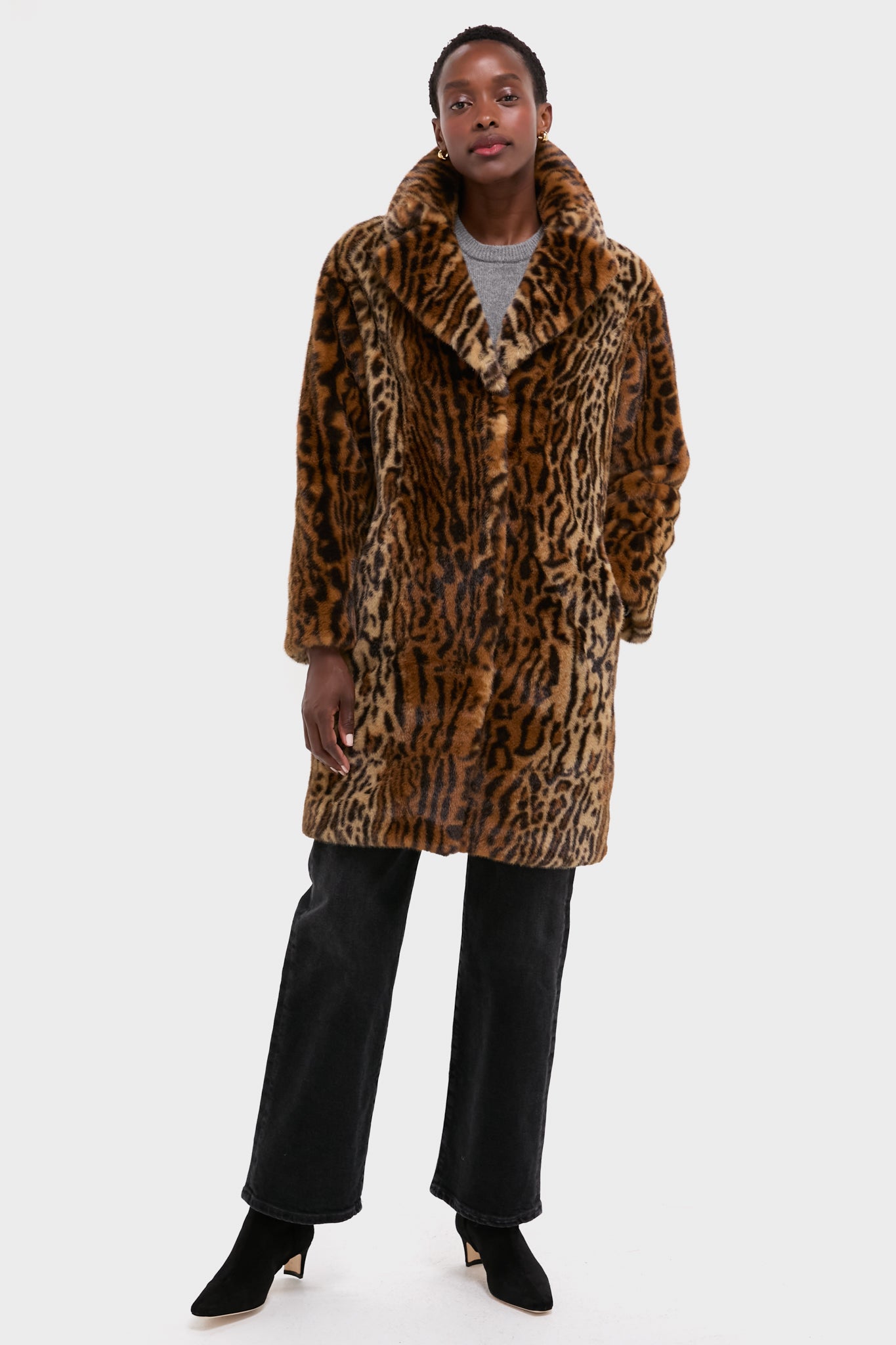 A person stands against a plain light background wearing a Stand Studio leopard print faux fur coat, gray sweater, black jeans, and black heeled boots.