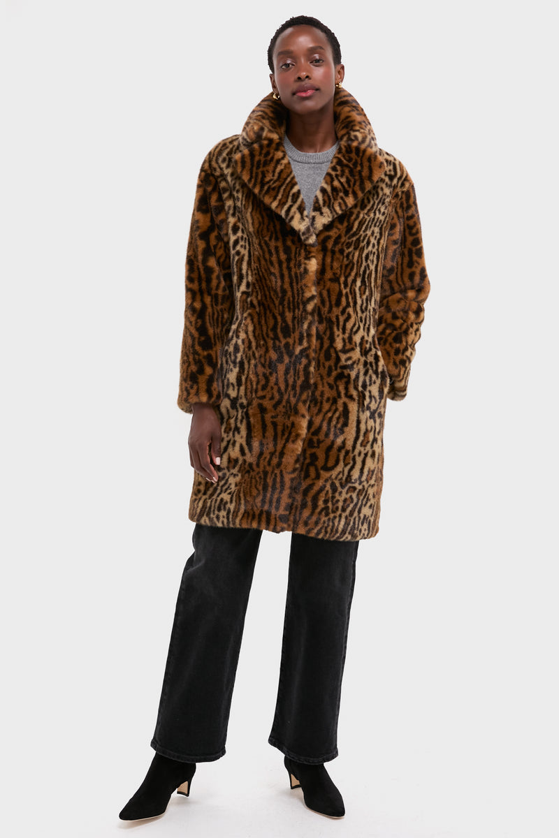A person stands against a plain light background wearing a Stand Studio leopard print faux fur coat, gray sweater, black jeans, and black heeled boots.