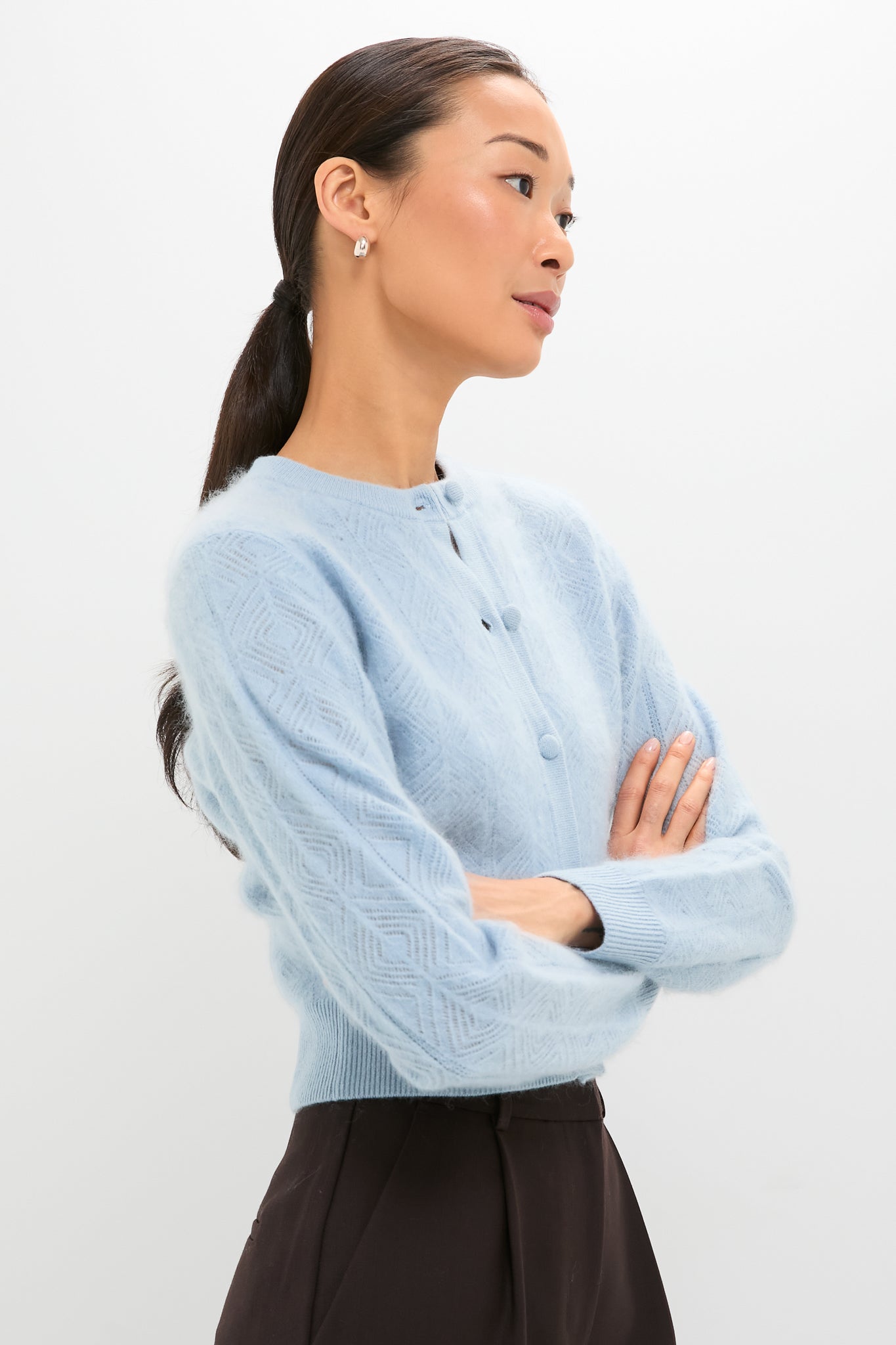 Model in profile wearing the Light Blue Brushed Merino Wool Macy Cardigan, featuring a classic diamond pattern and covered buttons, paired with dark high-waisted trousers for an effortless, high-end look.
