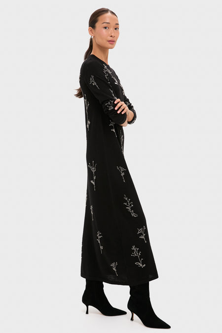 A woman stands sideways wearing an Altuzarra lama silk maxi dress with white floral patterns and beaded detailing, paired with black heeled boots against a plain light background.