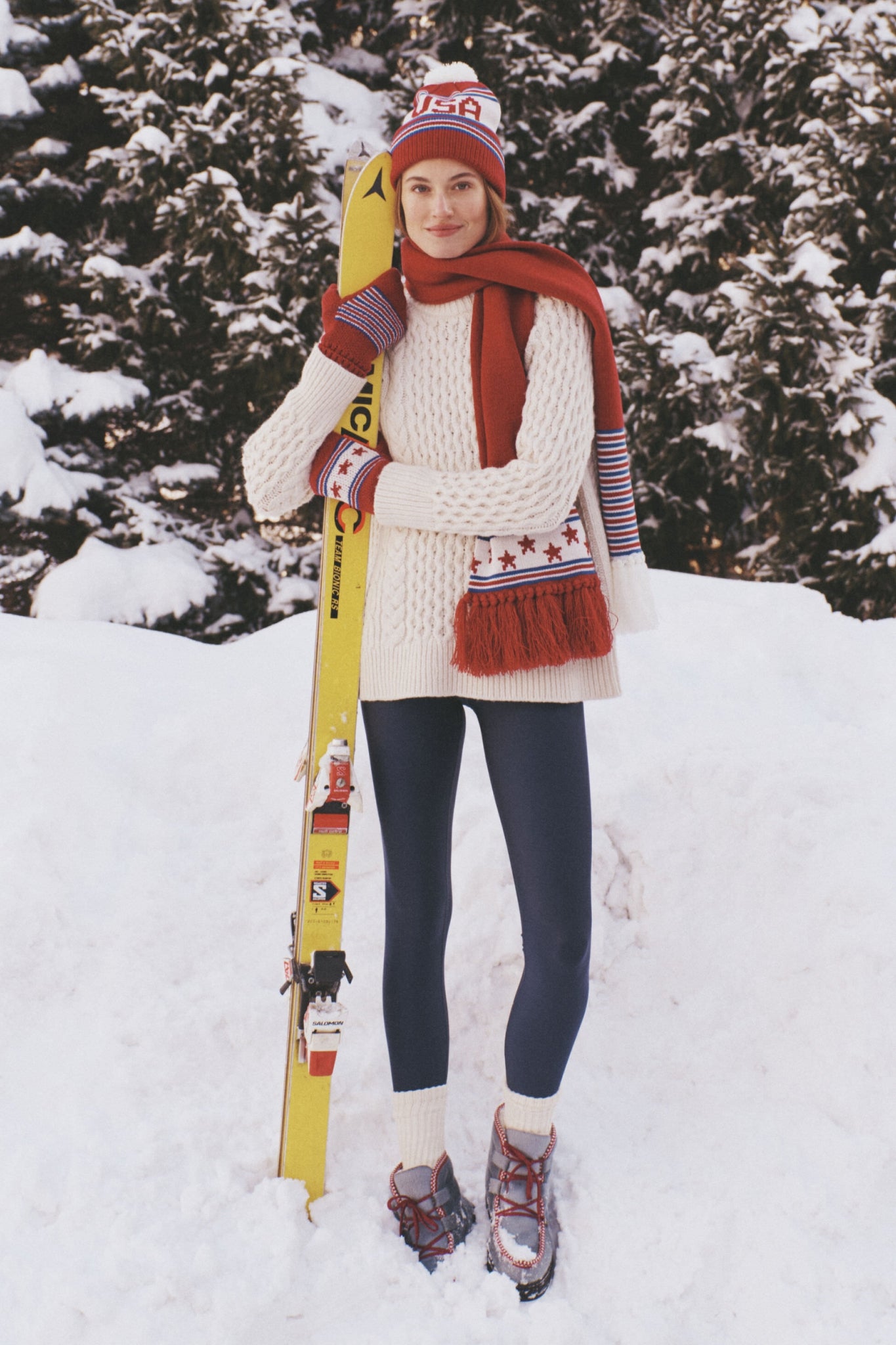 Person in snowy setting wearing the Ivory Galway Fisherman Sweater, showcasing its classic cable-knit design, effortlessly paired with winter sports gear and a bright yellow ski.