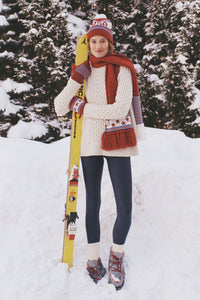Person in snowy setting wearing the Ivory Galway Fisherman Sweater, showcasing its classic cable-knit design, effortlessly paired with winter sports gear and a bright yellow ski.