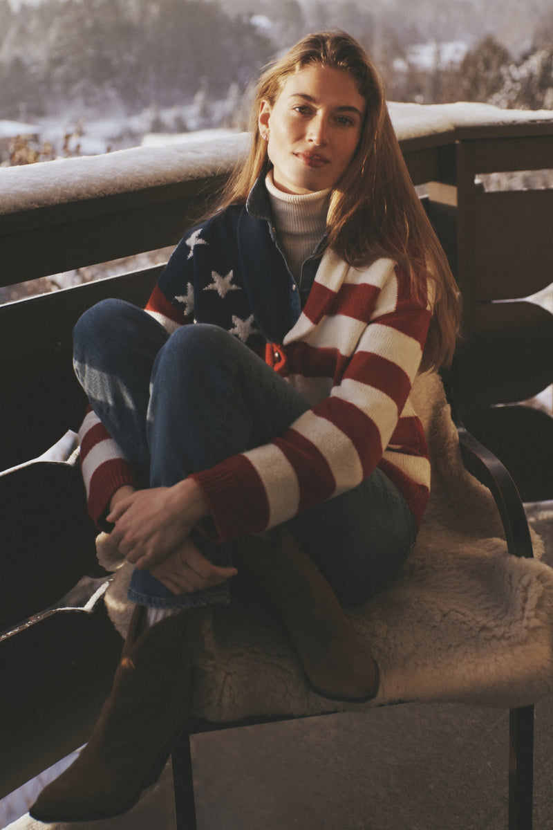 Young woman in classic American Flag Lindsey Cardigan on snowy porch, embodying effortless, high-end style with fun, sporty Americana vibes from Tuckernuck's winter collection.