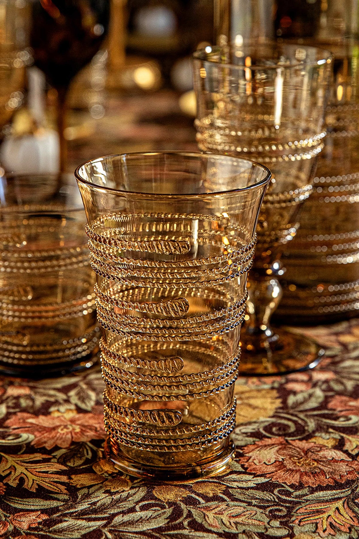 Amber Ella Large Tumbler with classic spiral design, mouth-blown, effortlessly combines fun and high-end style, set on an autumnal floral tablecloth with similar textured glassware behind.