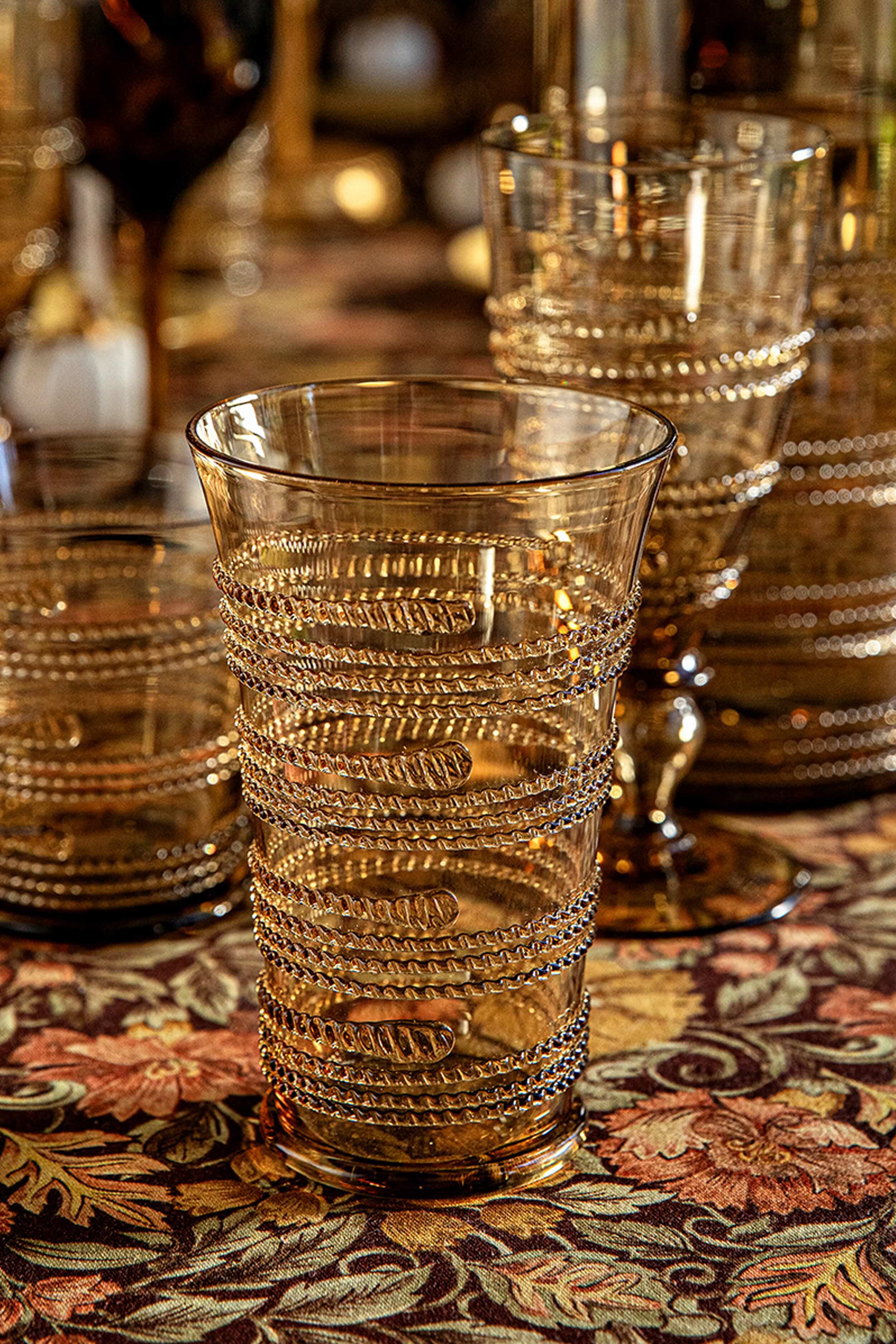 Amber Ella Large Tumbler with classic spiral design, mouth-blown, effortlessly combines fun and high-end style, set on an autumnal floral tablecloth with similar textured glassware behind.