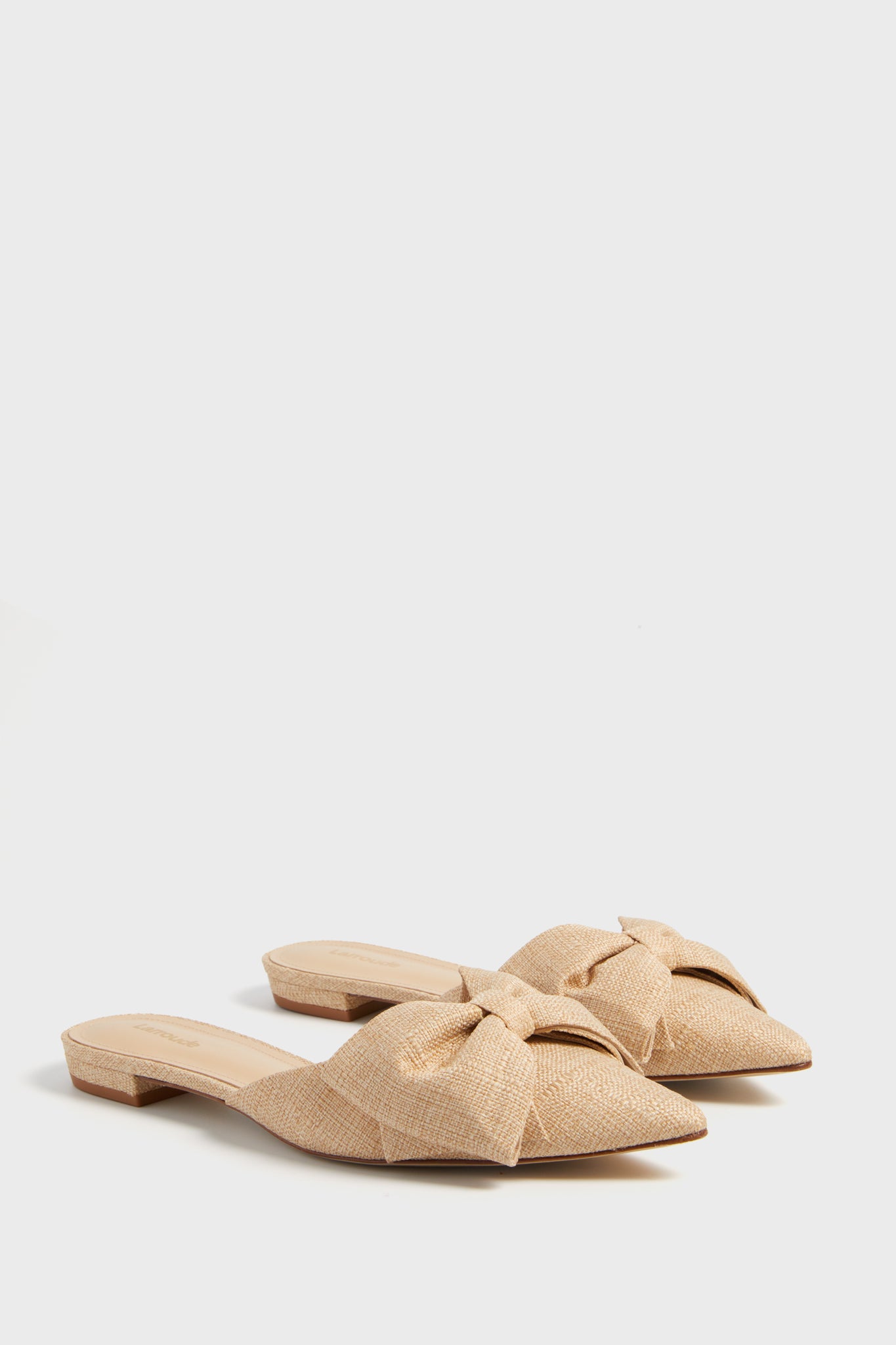 A pair of Larroude Elle Flat mules in beige raffia, featuring pointed toes and oversized bow accents on the upper, displayed in a minimalist setting.