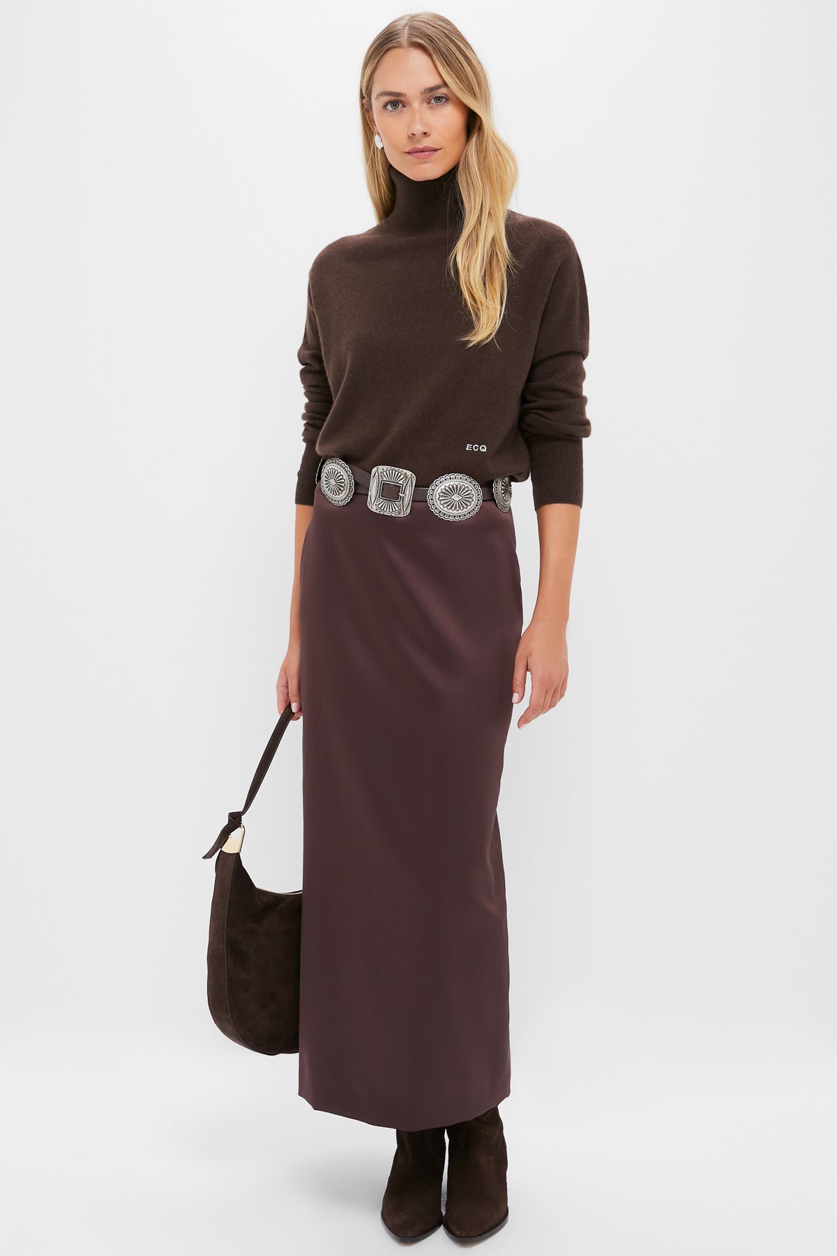 A woman in a Tuckernuck cashmere turtleneck sweater, dark brown long skirt, wide decorative belt, dark boots, and holding a brown bag stands against a plain white background.