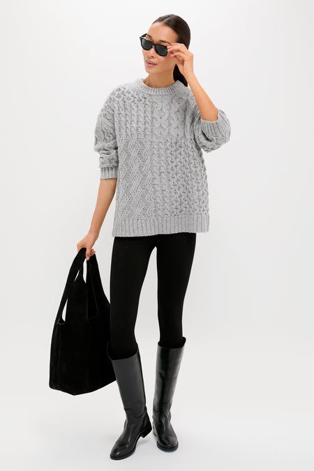 A person wearing a Tuckernuck grey cable-knit sweater with a relaxed fit pairs it with black leggings, knee-high boots, sunglasses, and a black tote bag, standing against a plain white background.