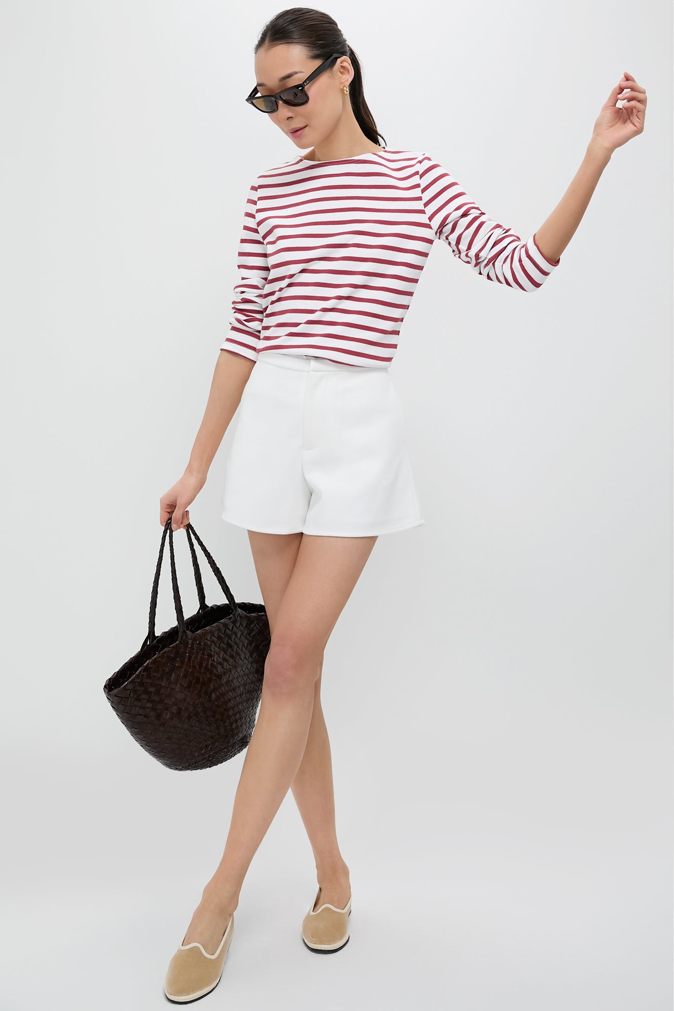 A woman in sunglasses wears a red and white striped nautical shirt, white shorts, tan slip-on flats, and carries a large black tote.