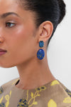 Close-up of a woman wearing Jennifer Behr Lapis Xiomara Earrings—striking 18K gold plated pieces with blue rectangular and oval stones—paired with handmade luxury accessories, hair pulled back, and a patterned top.