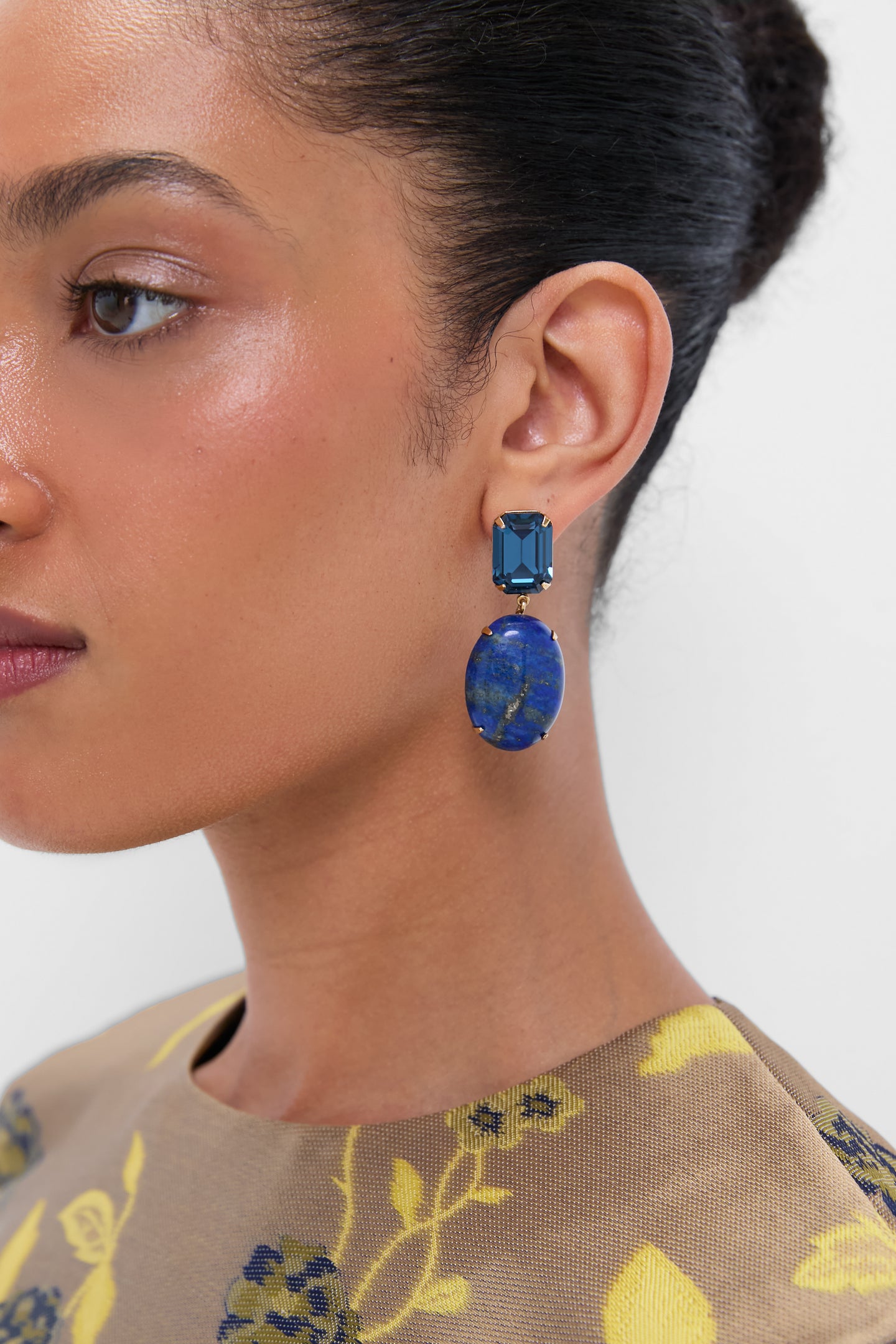 Close-up of a woman wearing Jennifer Behr Lapis Xiomara Earrings—striking 18K gold plated pieces with blue rectangular and oval stones—paired with handmade luxury accessories, hair pulled back, and a patterned top.