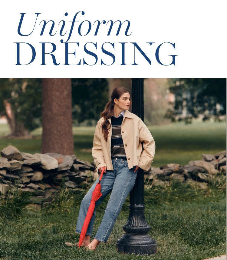 Woman leaning on a light post in a cropped trench coat, striped sweater, and jeans. Copy says: uniform dressing. 